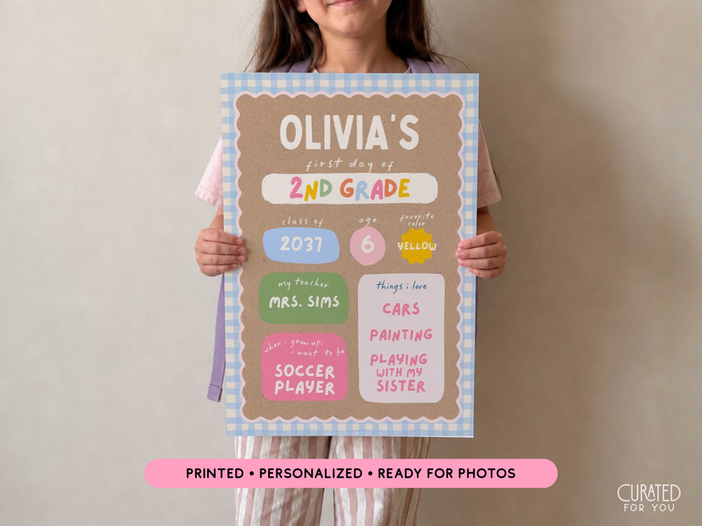 A child holds a personalized first-day-of-school sign that reads 'Olivia's first day of 2nd Grade' with pastel color-coded sections and decorative border. The child is partially visible behind the sign against a plain beige wall, with a pink banner at the bottom reading 'Printed • Personalized • Ready for Photos' and a 'Curated For You' mark in the corner.
Child holding a personalized 13 x 19 first day of school sign for back-to-school photos