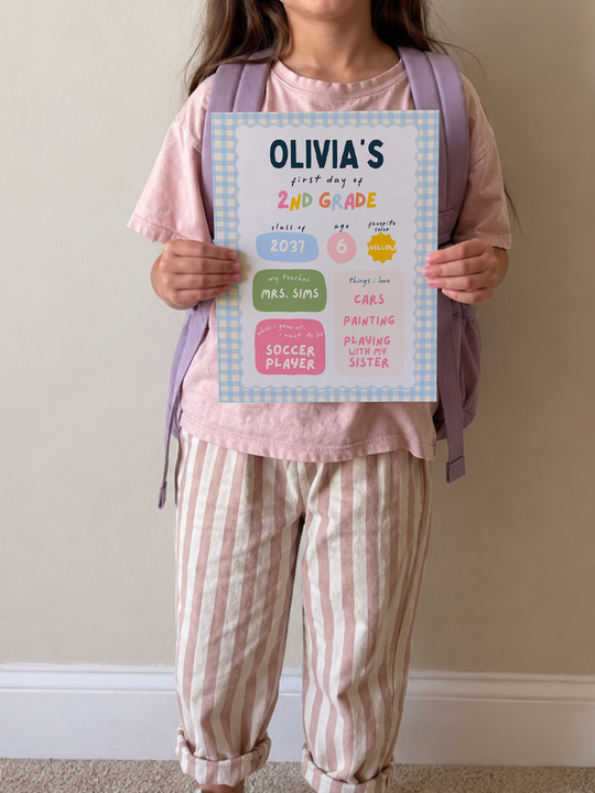 A young girl wearing a pink t-shirt and pink-and-white striped pants holds up a personalized first-day-of-school poster that reads &