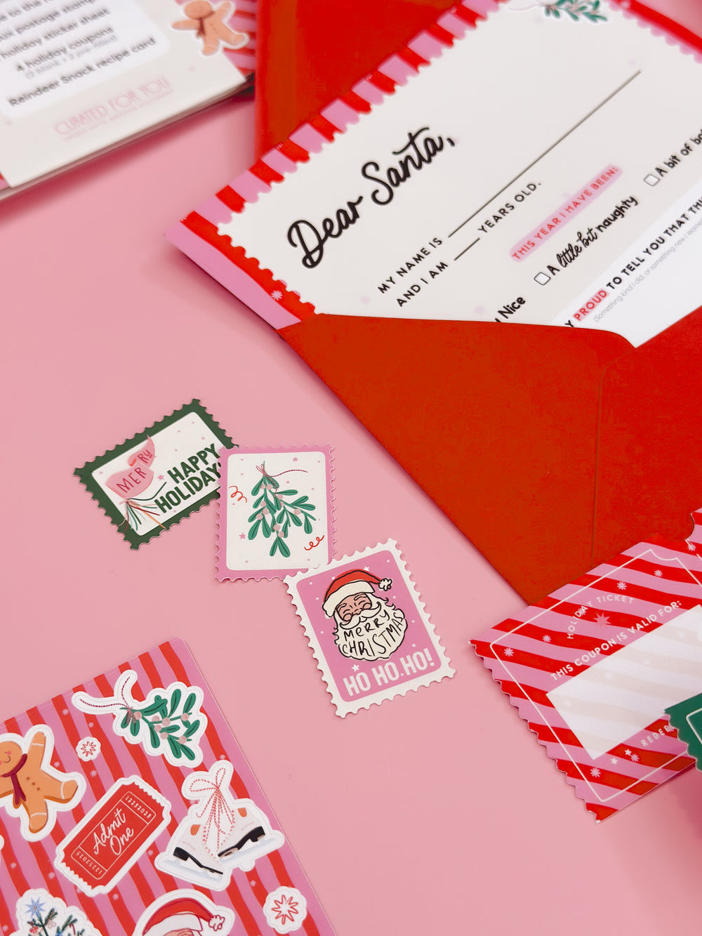 Christmas-themed stationery items including a letter to Santa and decorative stamps on a pink surface. Letter to Santa kit for kids with envelope, stamps, stickers, and holiday coupons.