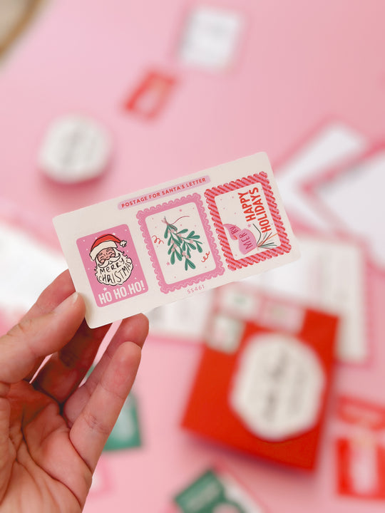 Hand holding a Christmas-themed postcard with decorative stamps on a pink background